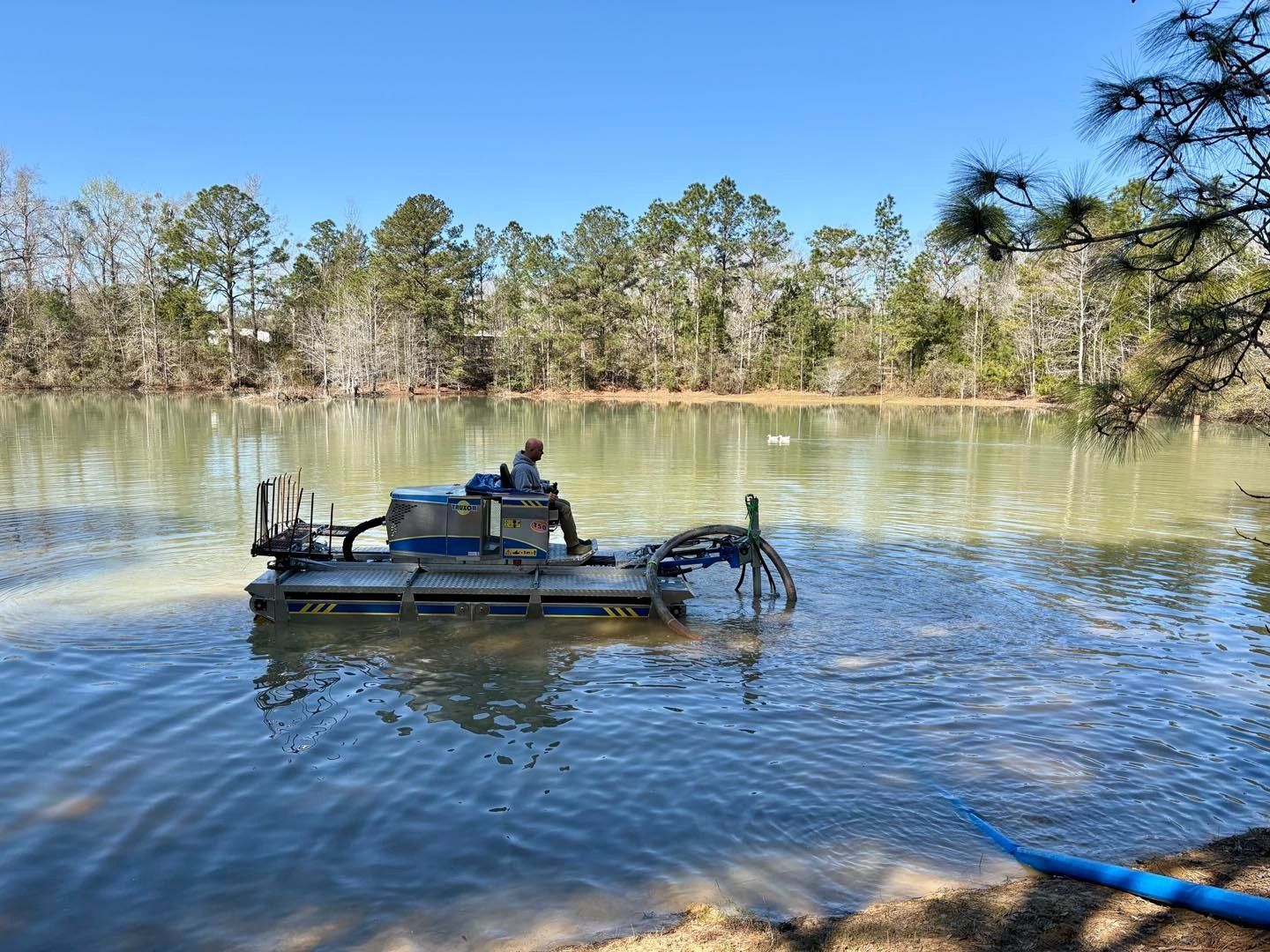 A person operates a blue machine in a murky pond to clean it, with trees in the background.