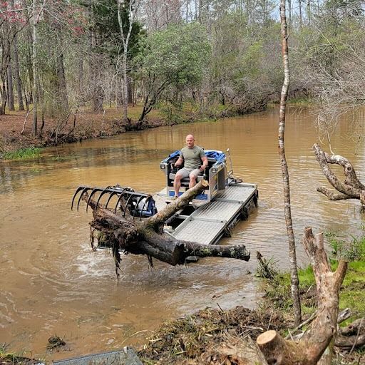 Man driving a machine with rake in murky water, clearing debris from a river. Outdoors setting.