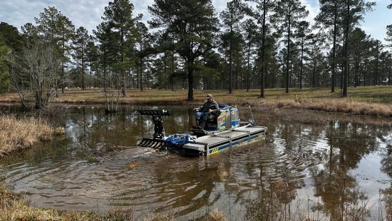 A person on a motorized boat in a pond, clearing weeds. Trees line the background.
