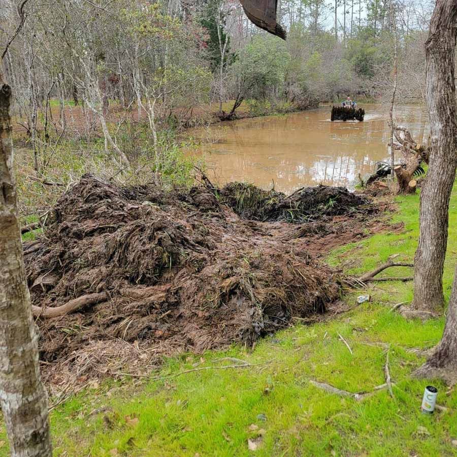 Pile of dirt and debris on green grass, pond in background with animal, trees.
