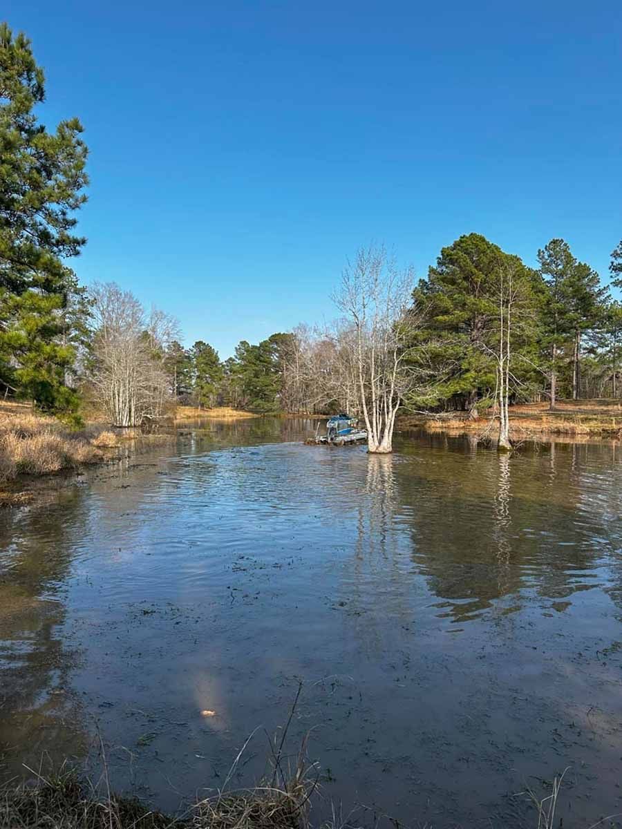 Pond reflecting a bright blue sky; bare and evergreen trees surround. A truck is stuck in the pond.