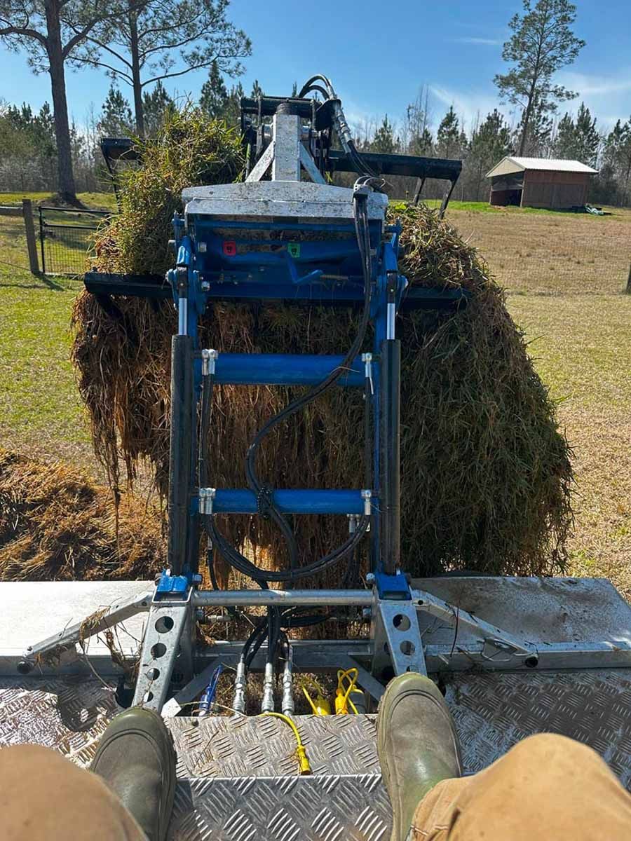 Person's view of a harvester collecting brown seaweed. Outdoors on a sunny day, with trees and a small building in the background.
