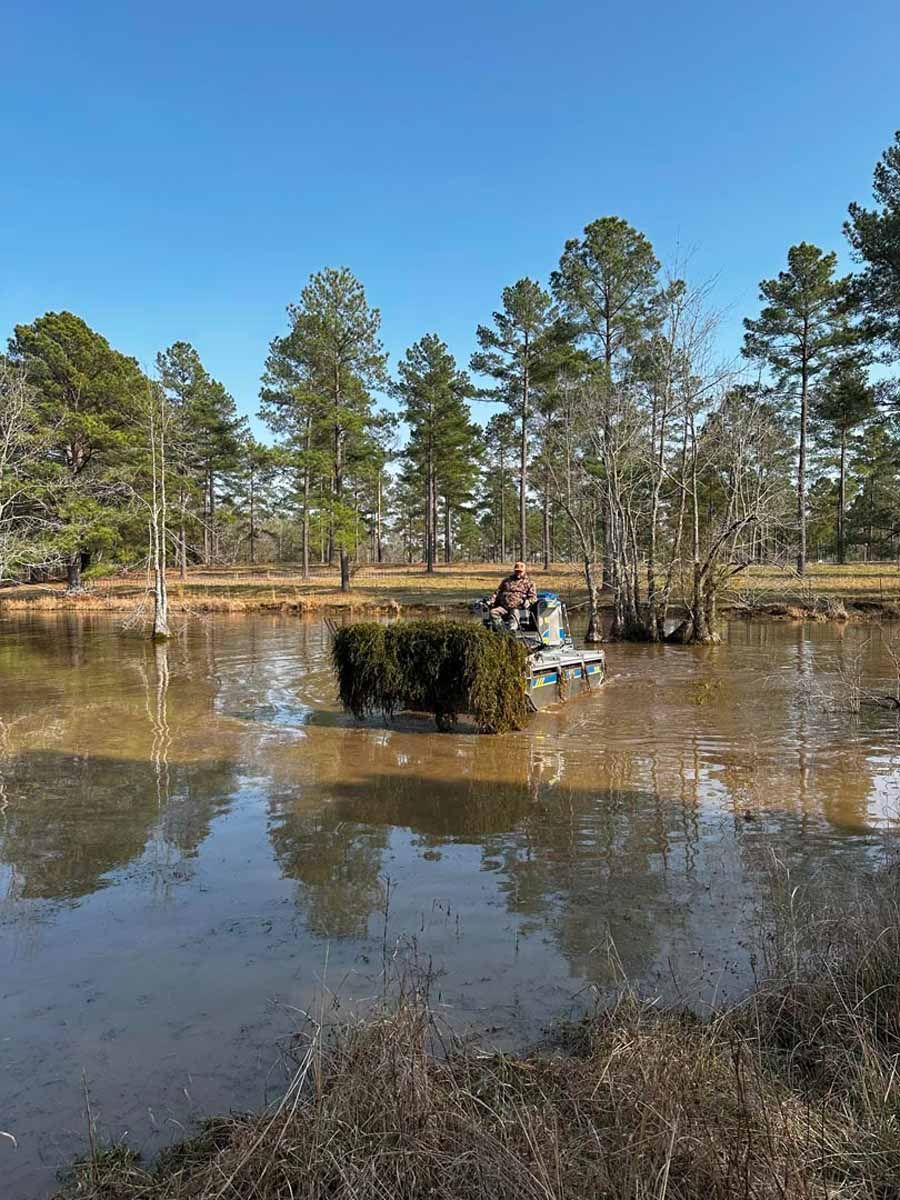 A person on a small machine cutting aquatic vegetation in a murky pond under a blue sky.