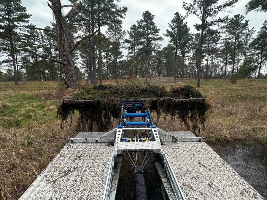 A specialized aquatic harvester collects vegetation in a wetland with trees.