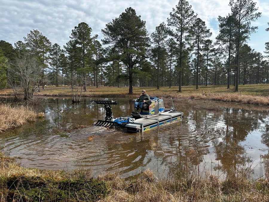 Man operating a blue aquatic weed harvester on a muddy pond. Trees and sky in the background.