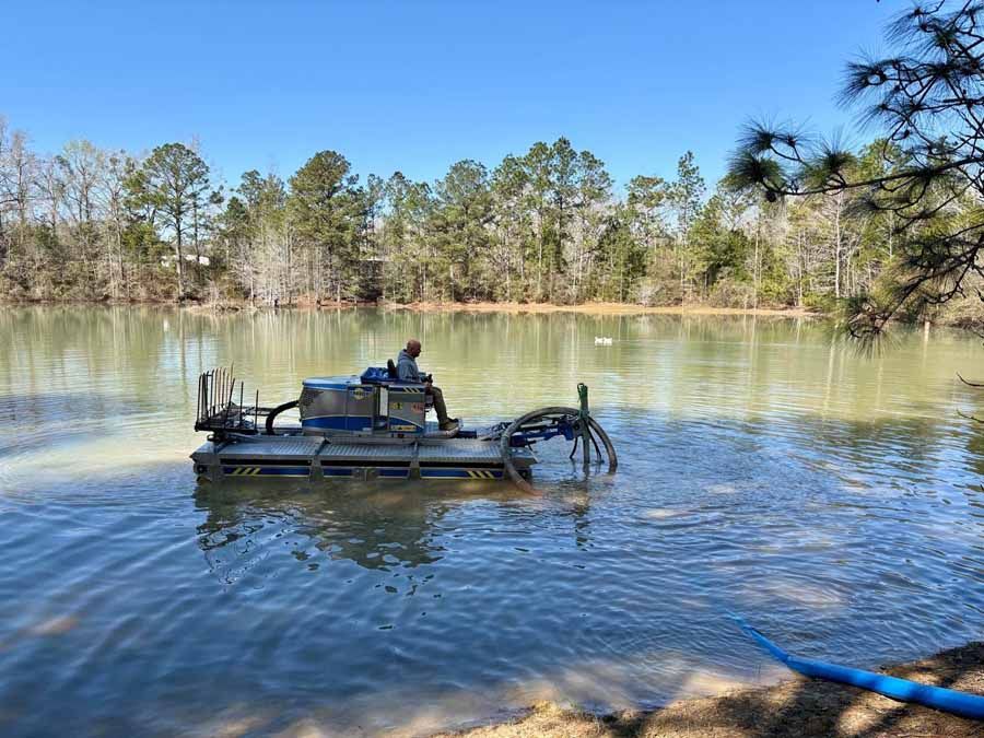 Man operating a blue dredge in a lake, removing sediment. Trees and blue sky in the background.