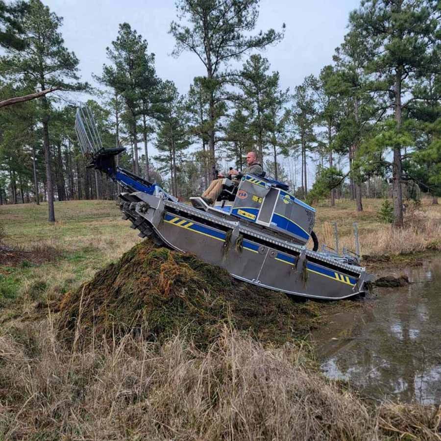 Person driving a metallic blue and gray amphibious machine on a bank of greenery near water, surrounded by trees.