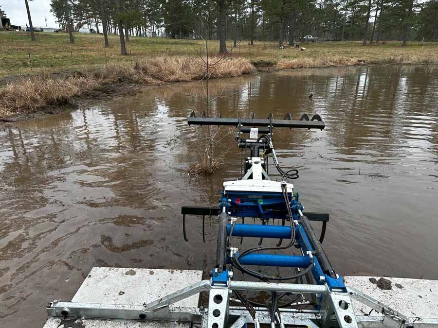A machine with rollers and blades in a murky pond, likely aerating or cleaning the water.