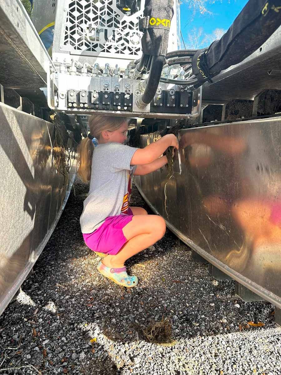 Girl in pink shorts and a gray shirt crouches, working on metal structure.