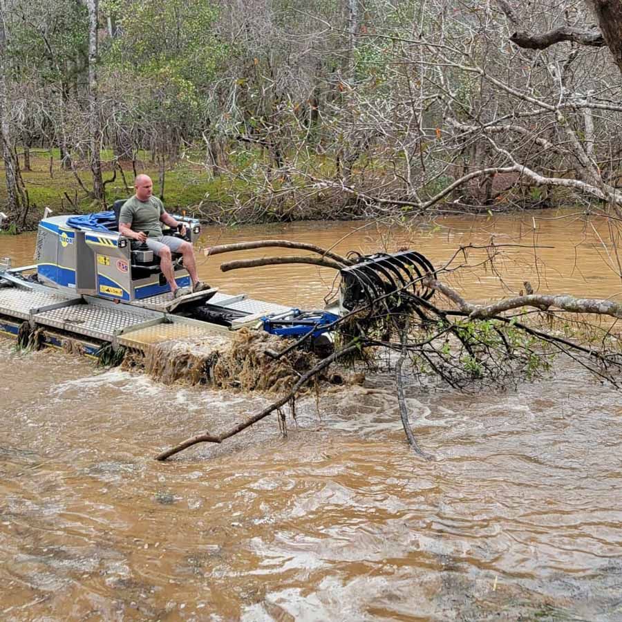 Man operating a watercraft with rake-like attachments, clearing debris from a murky river.