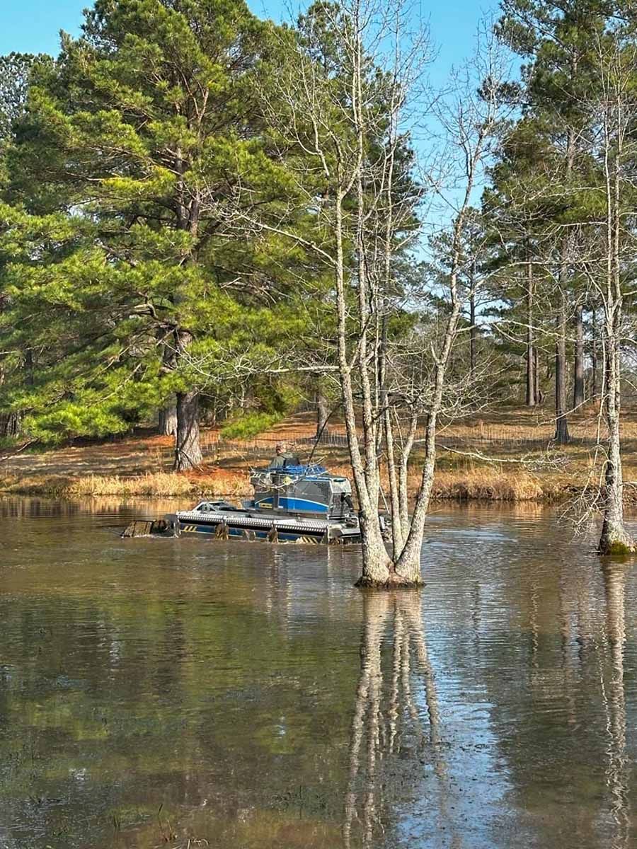 Trees and a machine sit in a reflective body of water, with a forest background.