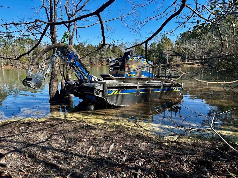 A blue and black excavator in a lake, clearing debris. Green trees and blue sky in the background.