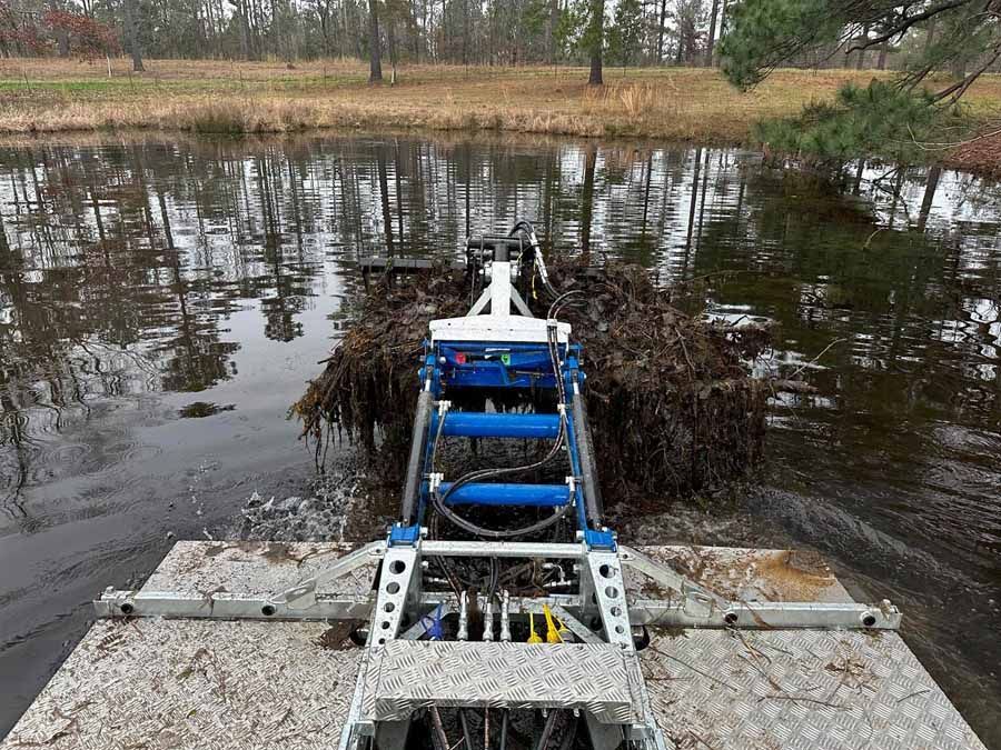A machine harvesting weeds from a pond; it has a blue and silver frame with a large clump of weeds.