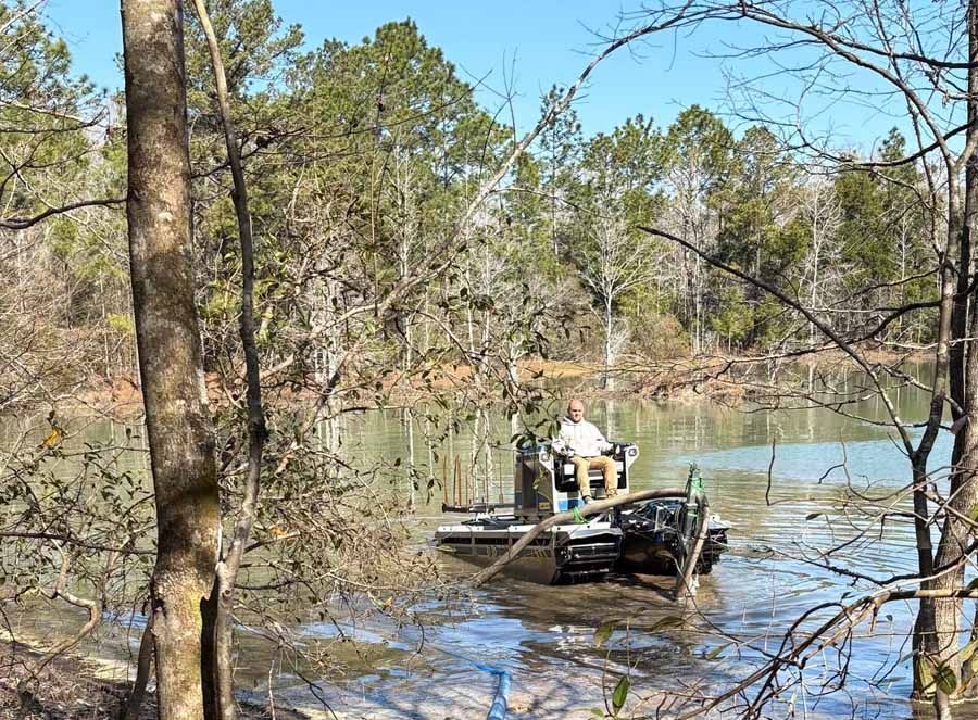 Man in a motorized wheelchair navigating a swamp. Brown water, green trees in background, sunny day.