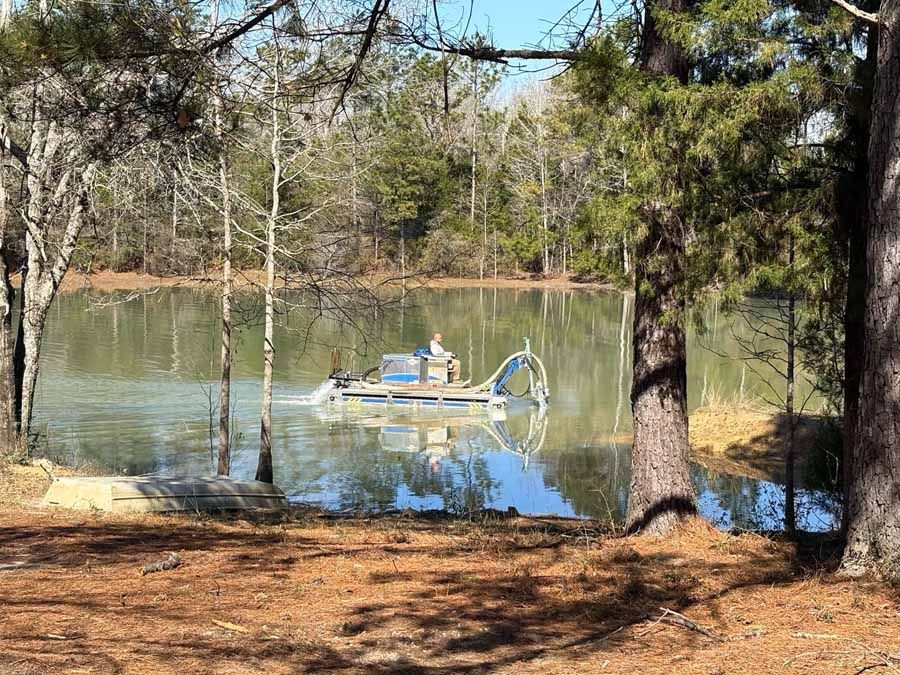 A person operates a machine on a pond. Trees surround the water.