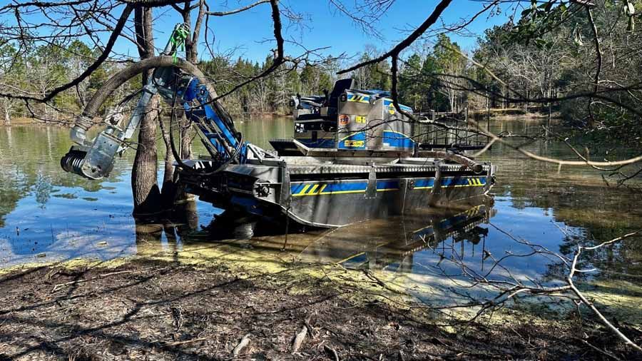 Floating excavator in a pond, removing debris; blue water, trees, and a bright sunny day.