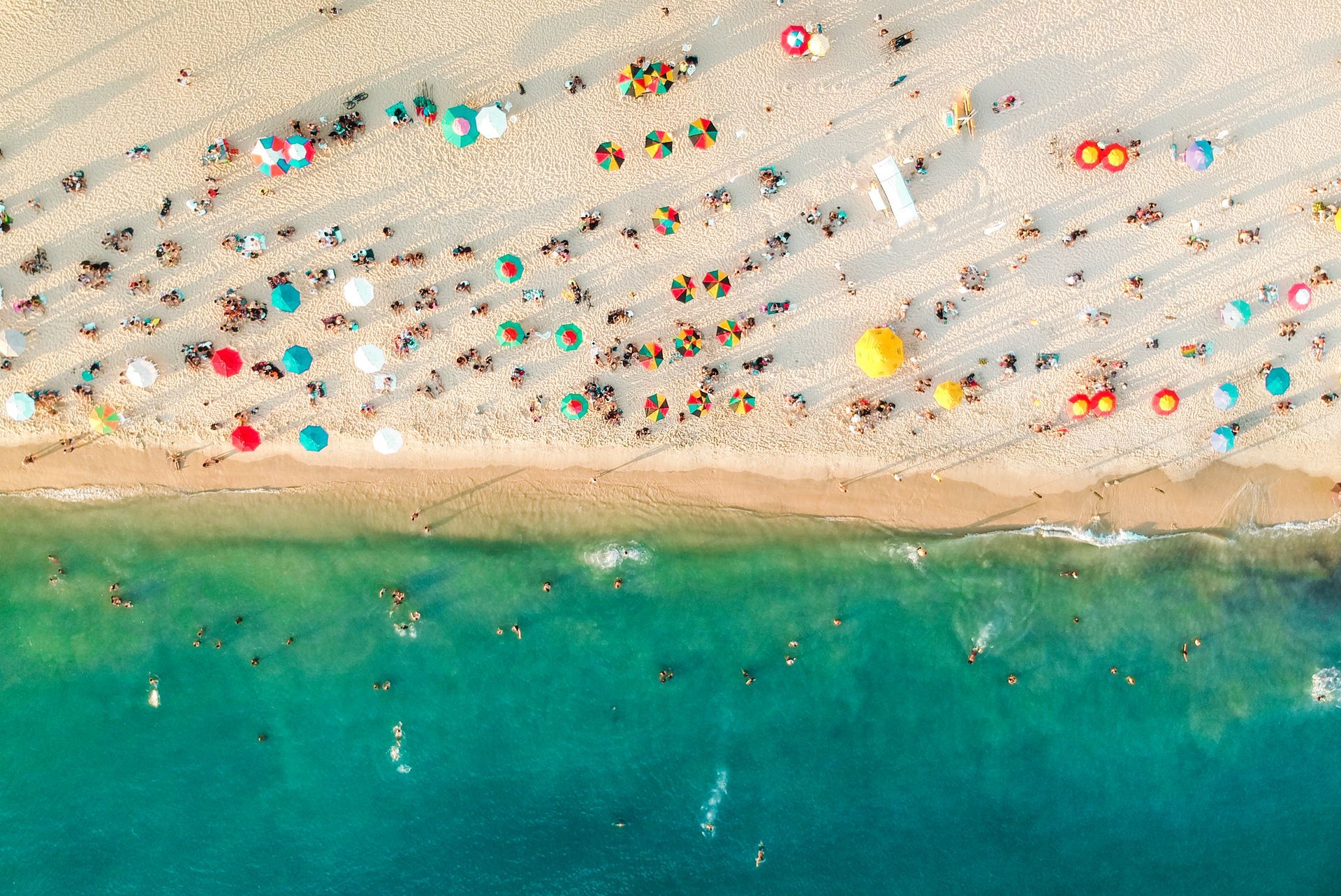 Aerial view of a crowded beach with colorful umbrellas and people swimming in turquoise water.