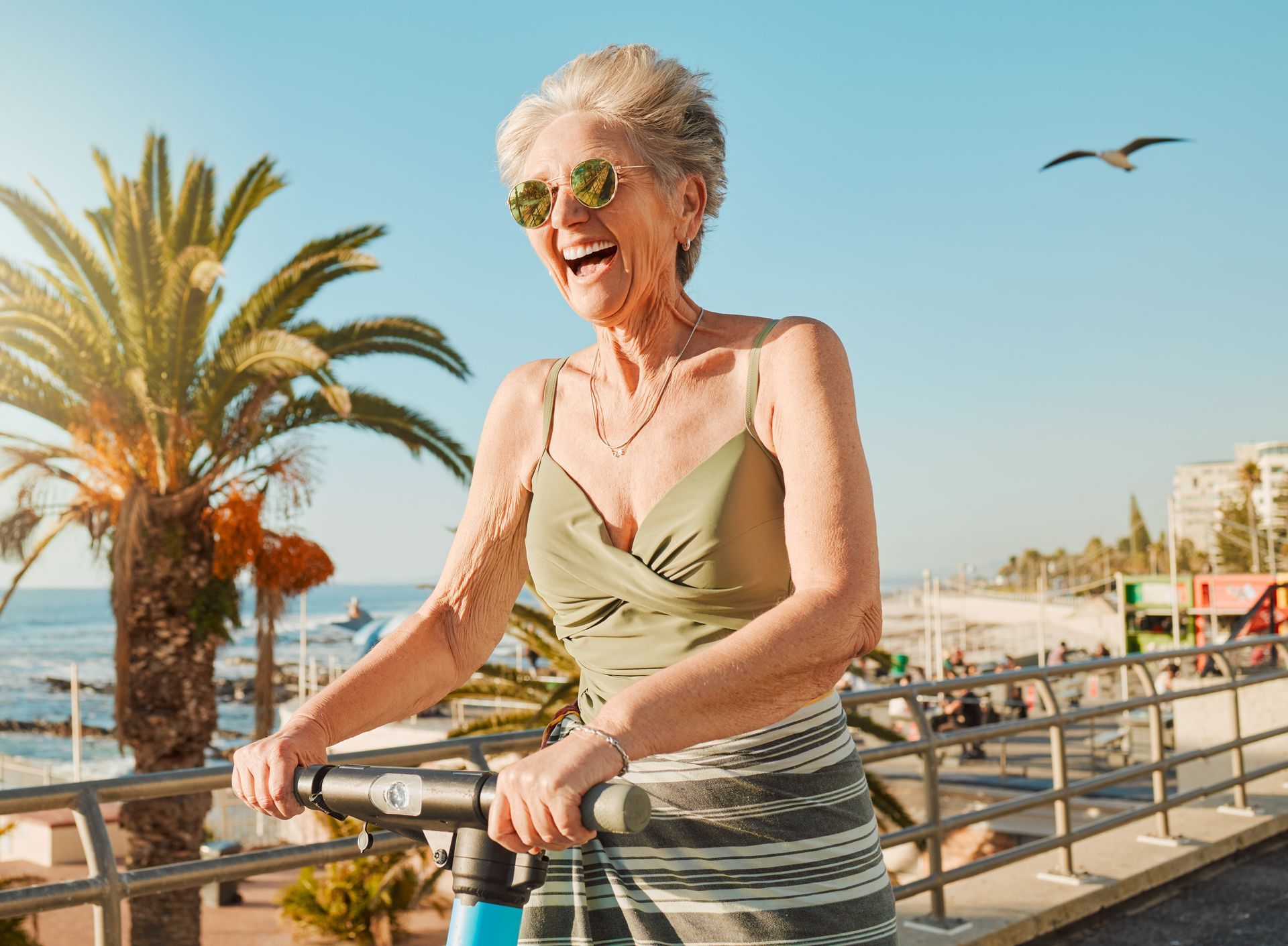Woman riding a scooter, laughing near a beach with palm trees, wearing sunglasses and a tank top.