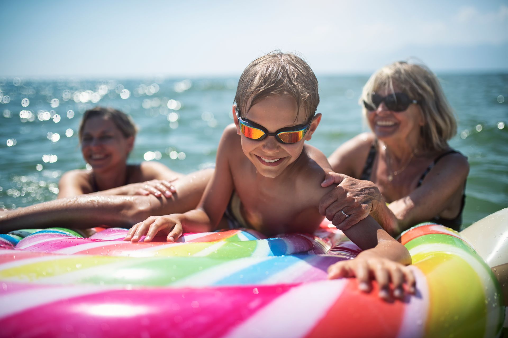 Boy wearing sunglasses smiles on a rainbow floatie in the ocean with two adults.