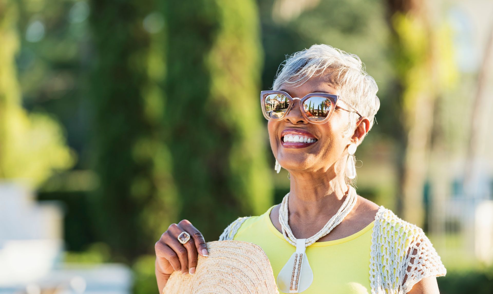 Woman in sunglasses smiling, holding a straw hat, wearing yellow top and pearl necklace.