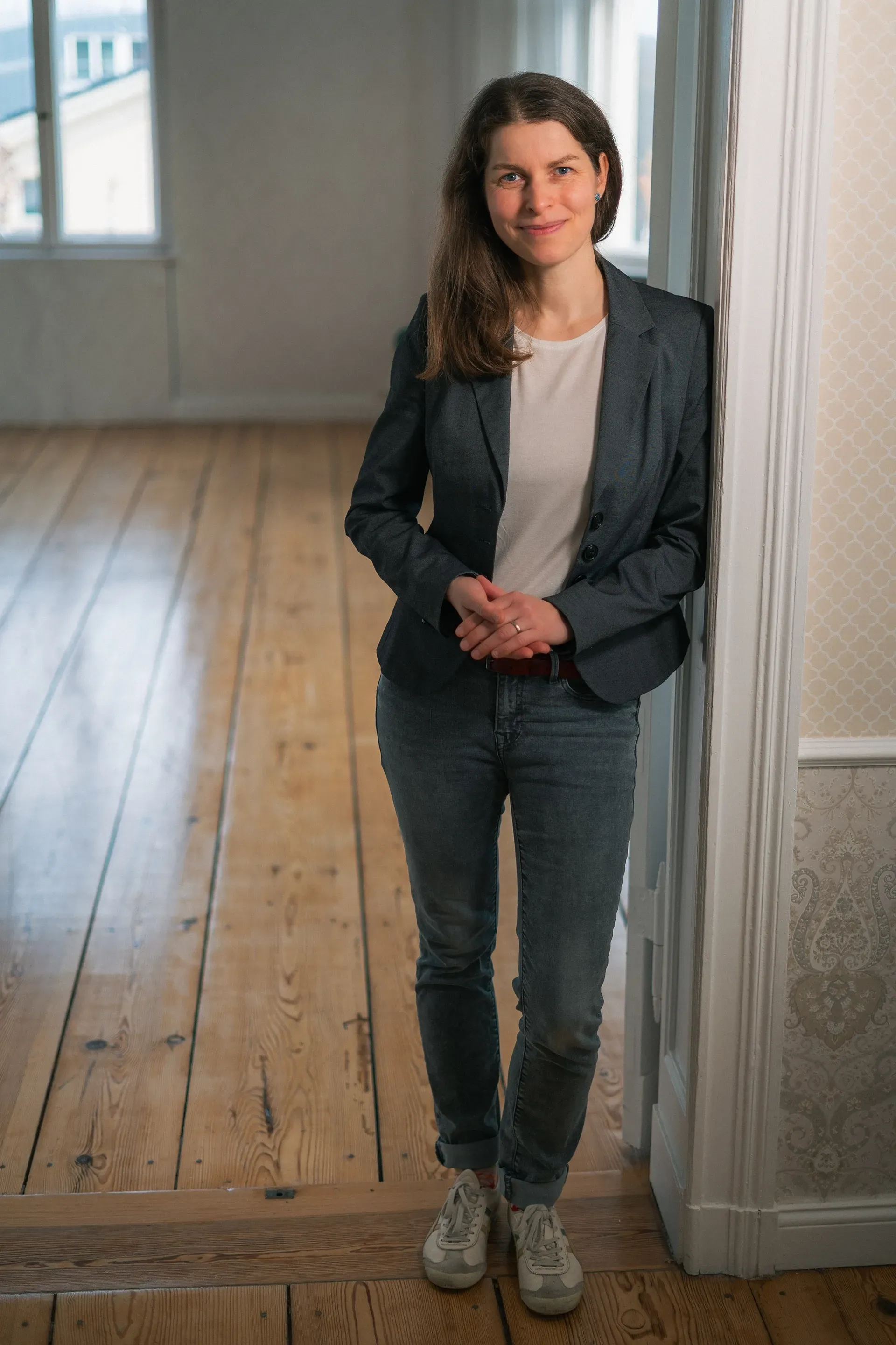 Antje in Blazer und Jeans lehnt in einem leeren Raum mit Holzboden an einem Türrahmen.