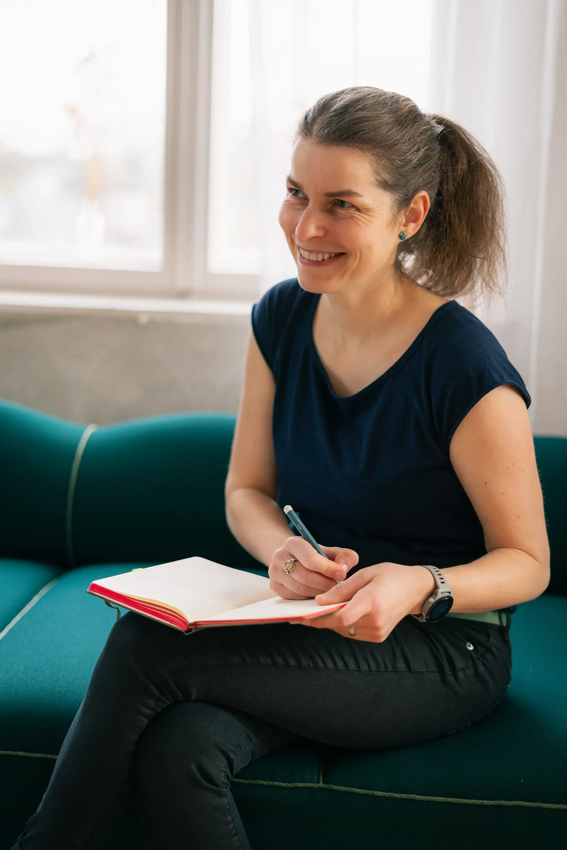 Eine lächelnde Frau schreibt in ein Notizbuch und sitzt auf einem türkisfarbenen Sofa am Fenster.