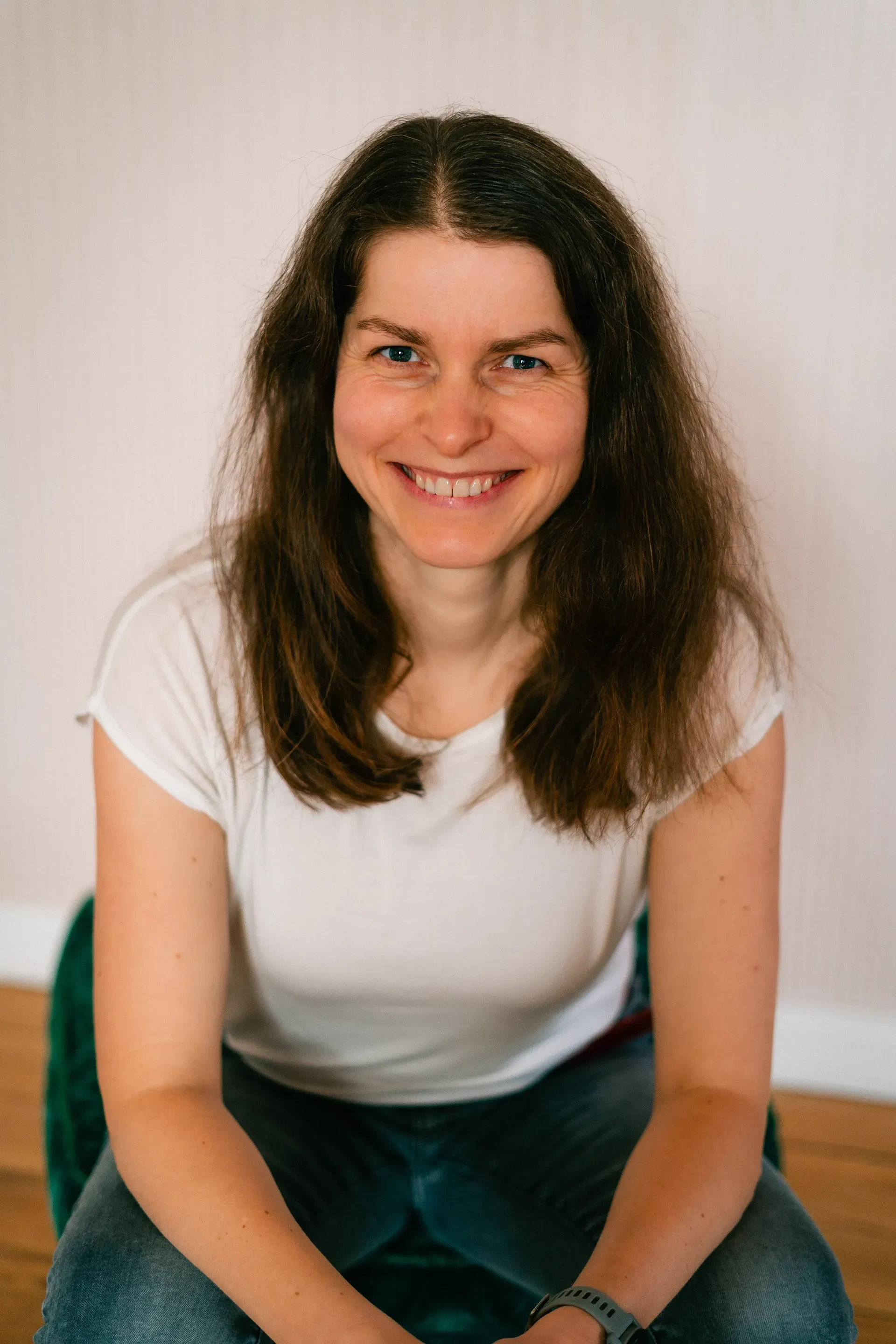 Eine Frau mit langen braunen Haaren lächelt, sitzt auf einem Stuhl und trägt ein weißes T-Shirt und Jeans. Sanftes, natürliches Licht.