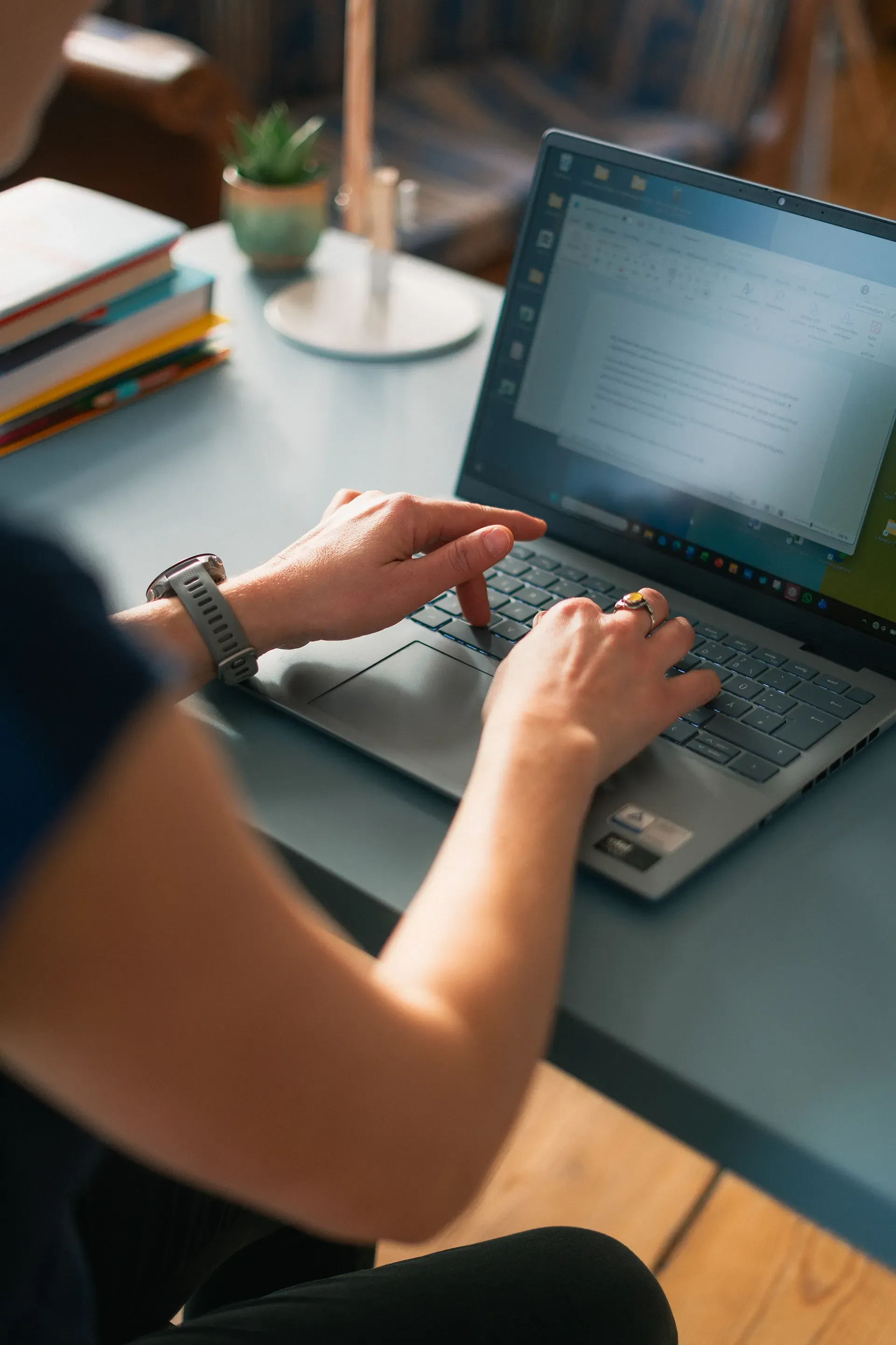 Eine Person tippt an einem Laptop an einem blauen Schreibtisch; in der Nähe befinden sich Bücher und eine Pflanze.