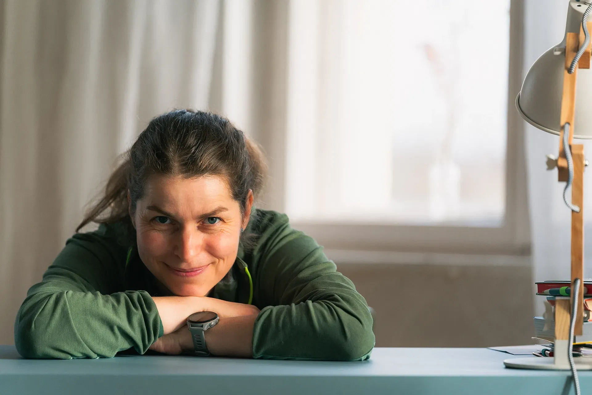 Antje stützt Kopf lächelnd an einem blauen Schreibtisch auf den Armen ab. Sie trägt ein grünes Hemd und eine Smartwatch und befindet sich drinnen am Fenster.