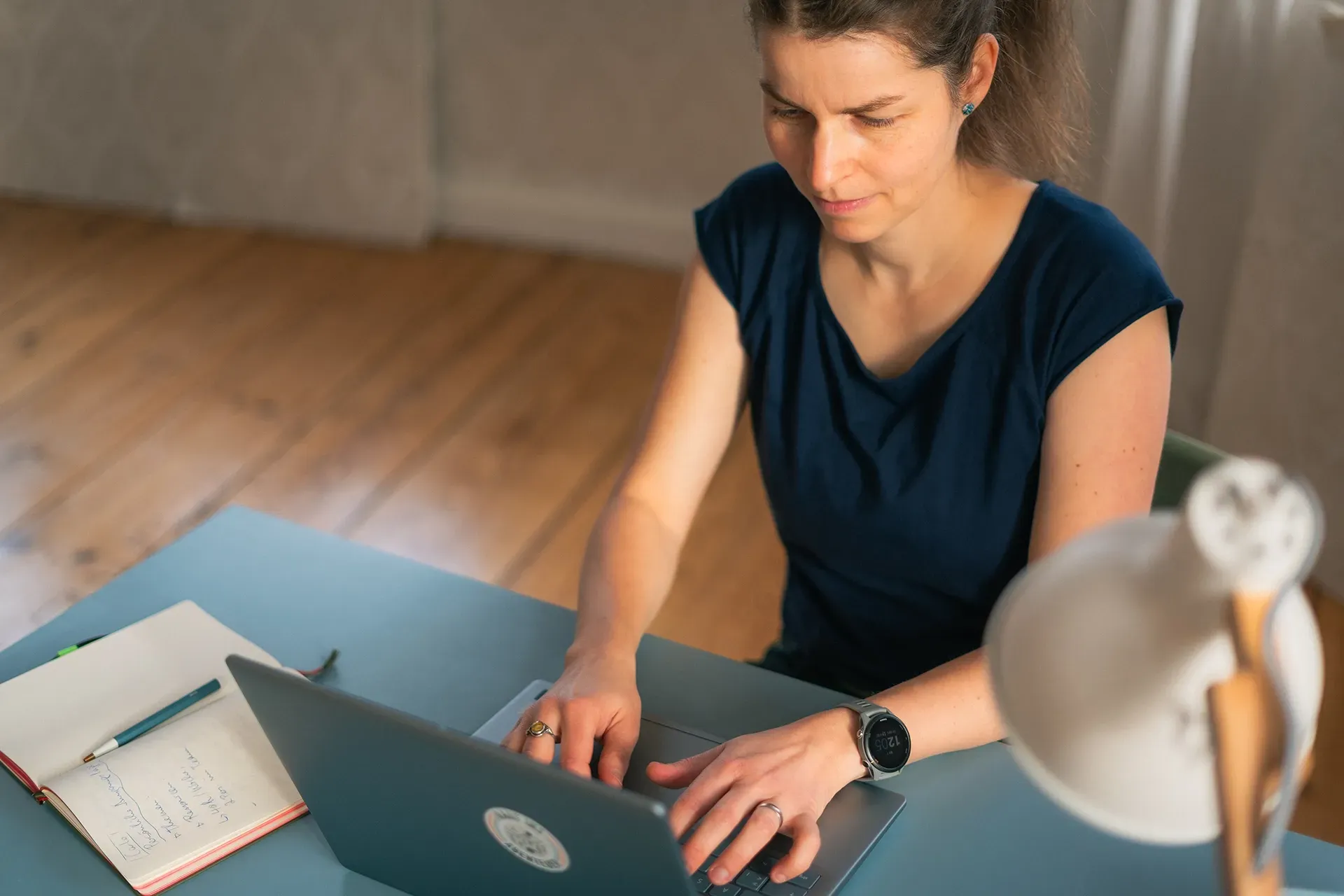 Antje tippt an einem blauen Schreibtisch auf einem Laptop; Notizbuch und Lampe sind zu sehen.
