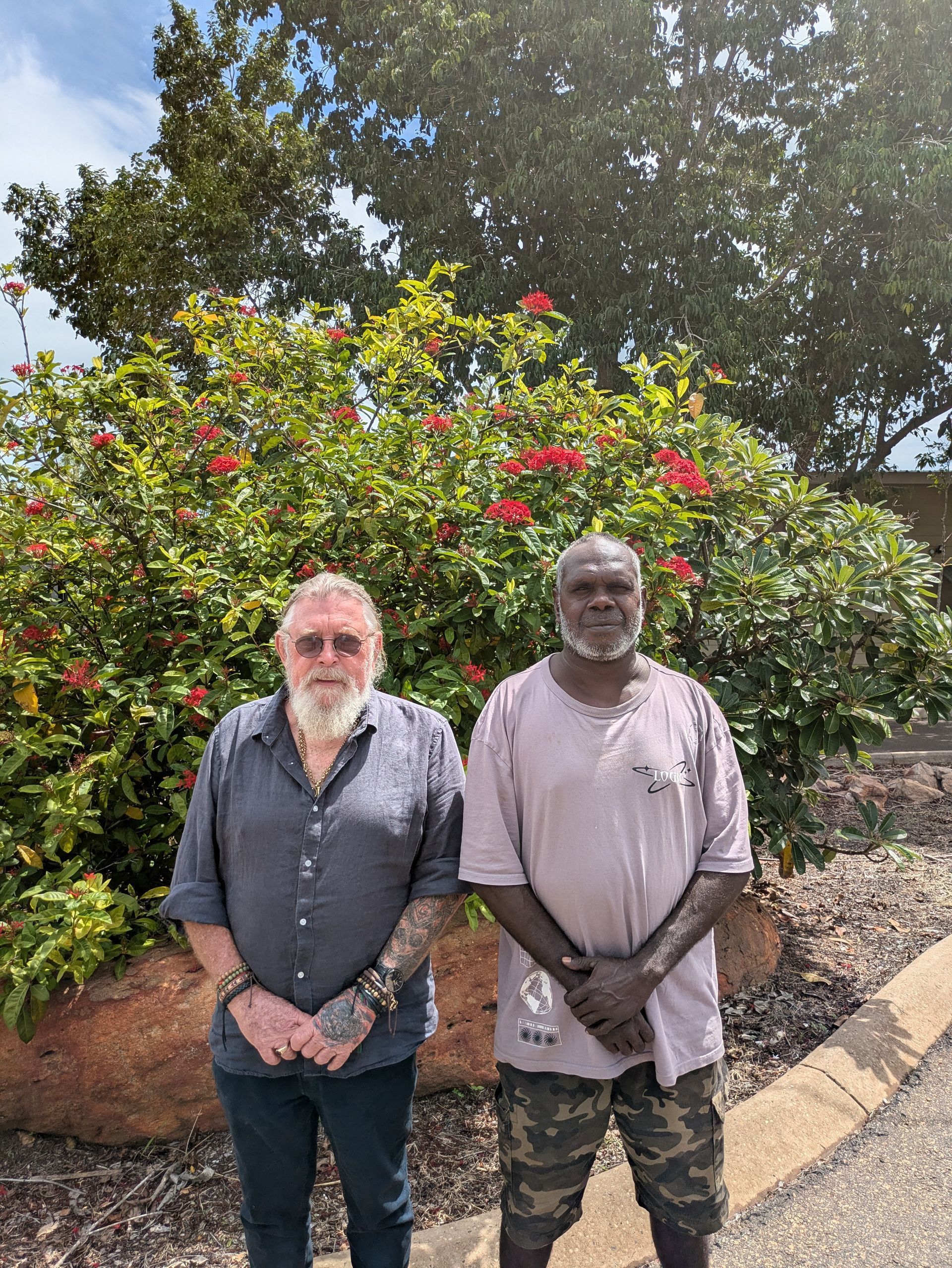 Two men standing in front of a bush with red flowers. One has a beard, the other is in camouflage.