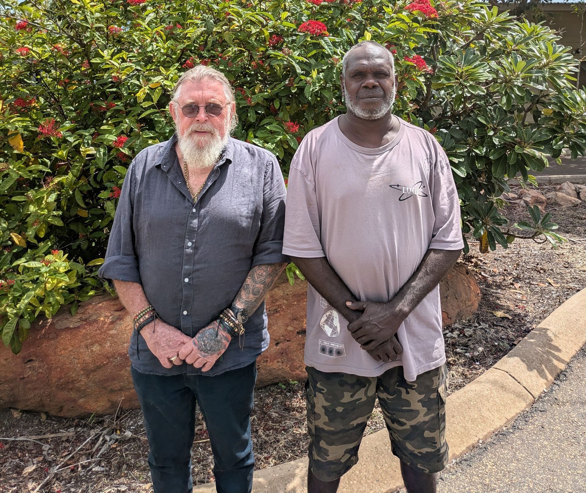 Two men stand outside by red flowering plants. One has a beard and tattoos; the other wears camouflage shorts.