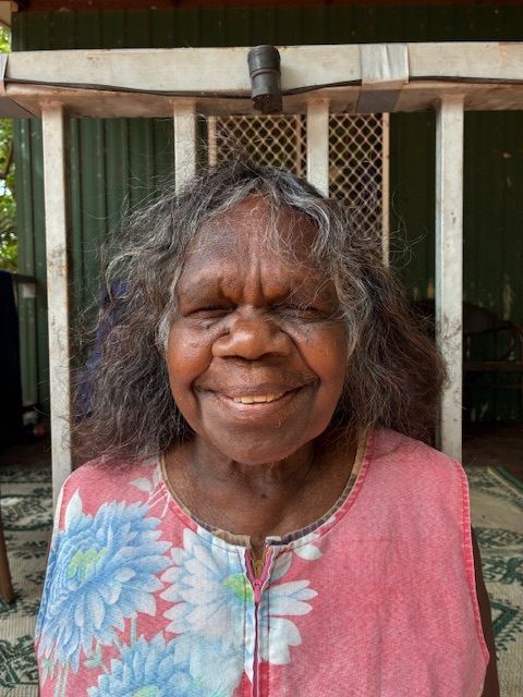 Elderly Indigenous woman smiling, wearing pink floral top, standing on a porch.