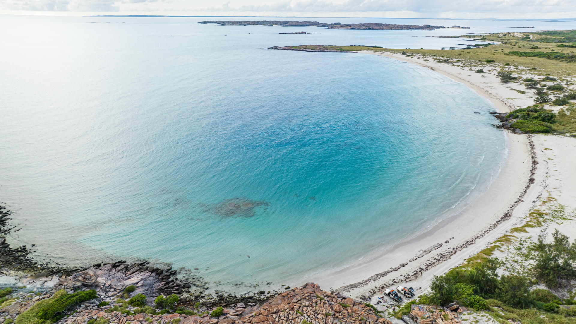 Aerial view of a pristine, crescent-shaped beach with clear turquoise water and white sand, surrounded by rocky and grassy land.
