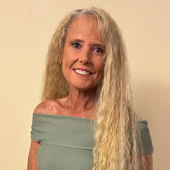 Woman with long blonde crimped hair, smiling, wearing an off-shoulder sage green top.