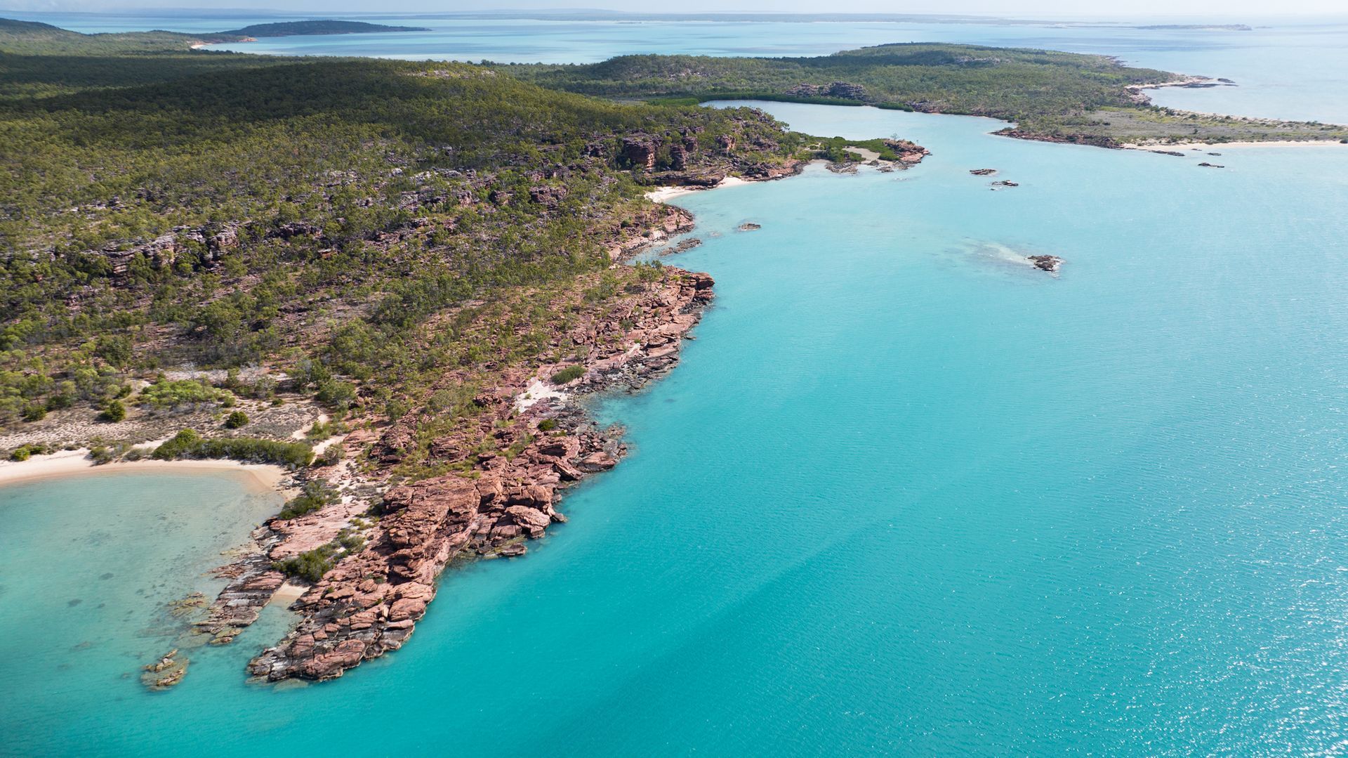 Turquoise water meets a rocky shoreline dotted with green trees under a bright sky.