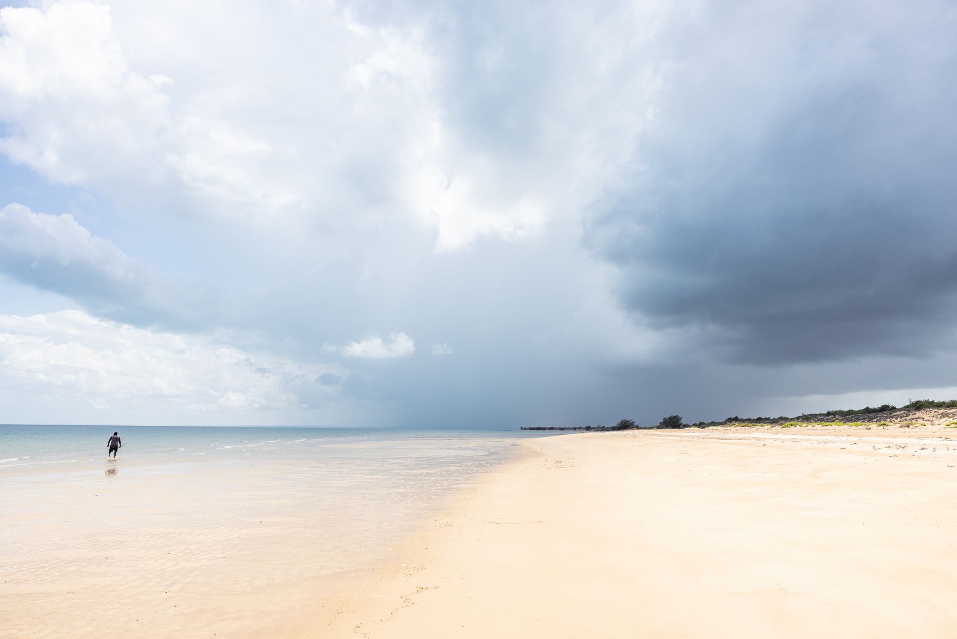 Beach scene under a stormy sky, a person wades in shallow water, sand and dark clouds dominate the scene.