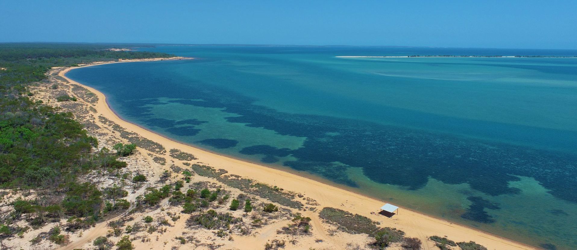 Aerial view of a beach with turquoise water, sandy shore, and a green coastal area under a clear blue sky.