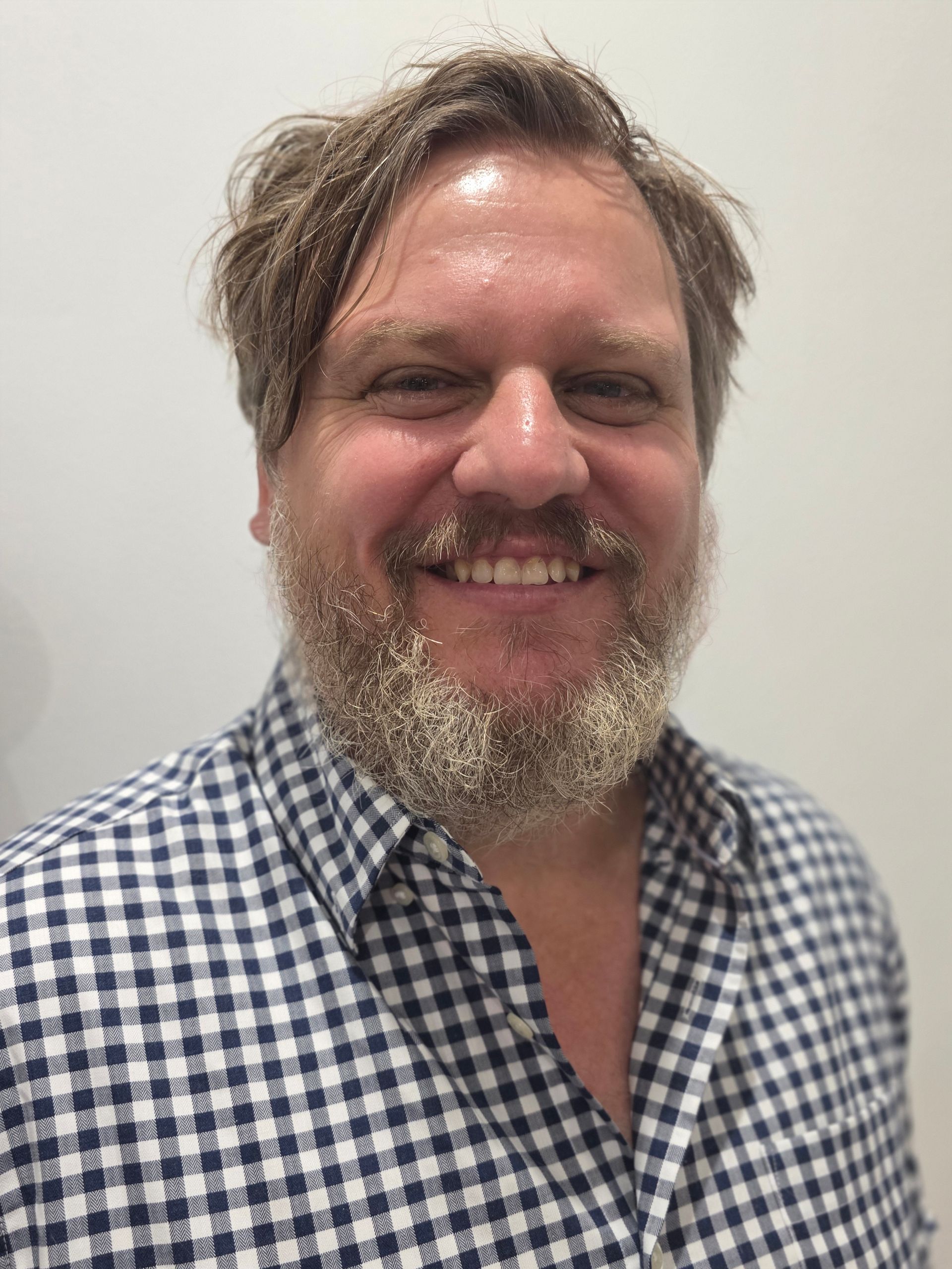 Man with beard smiles, wearing a blue and white checkered shirt.