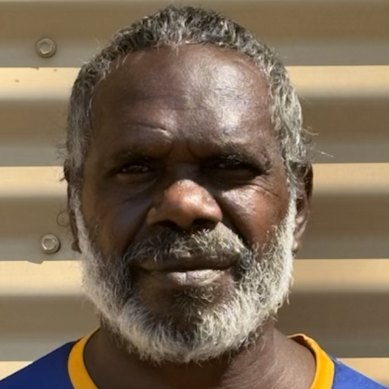 Man with grey beard and dark skin wearing a blue and yellow shirt, against a neutral background.
