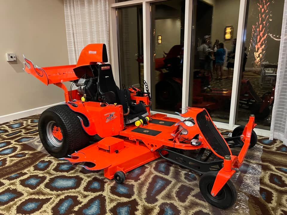 An orange lawn mower is parked in a room next to a window.