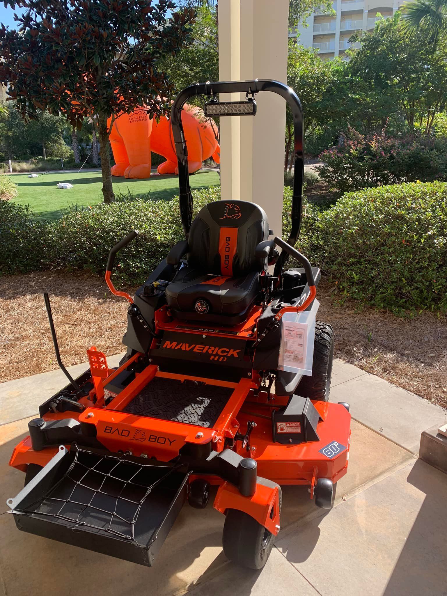 An orange and black lawn mower is parked on a sidewalk in front of a park.