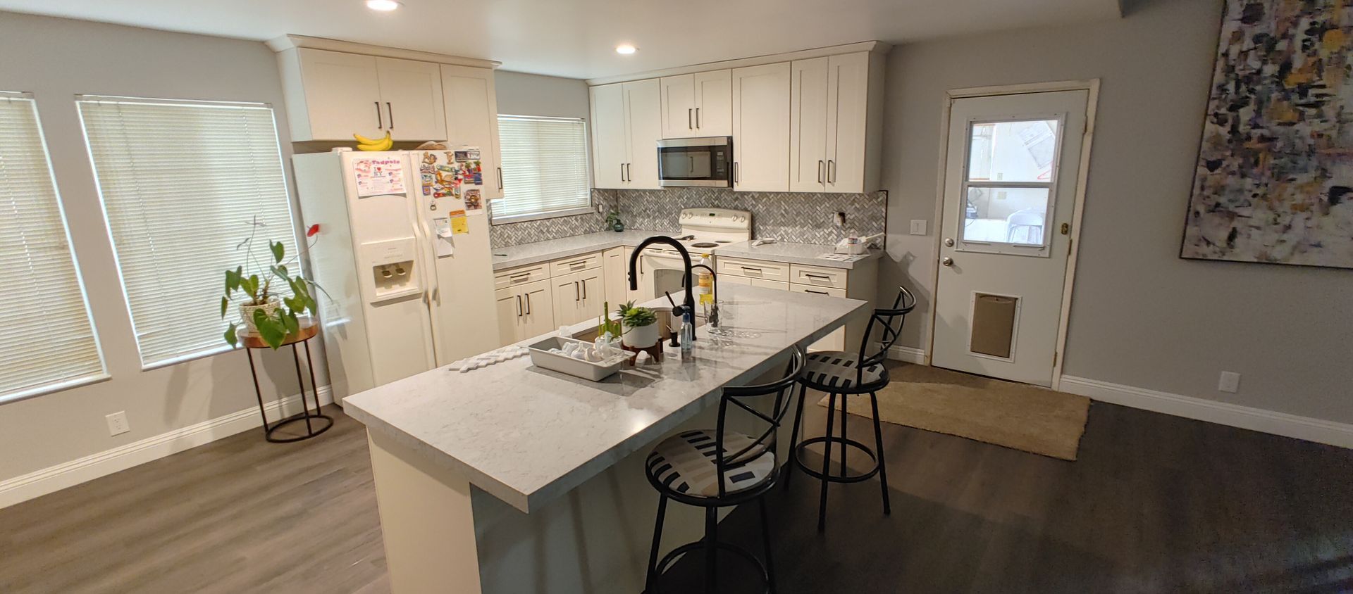 A kitchen with white cabinets , a large island , stools and a refrigerator.