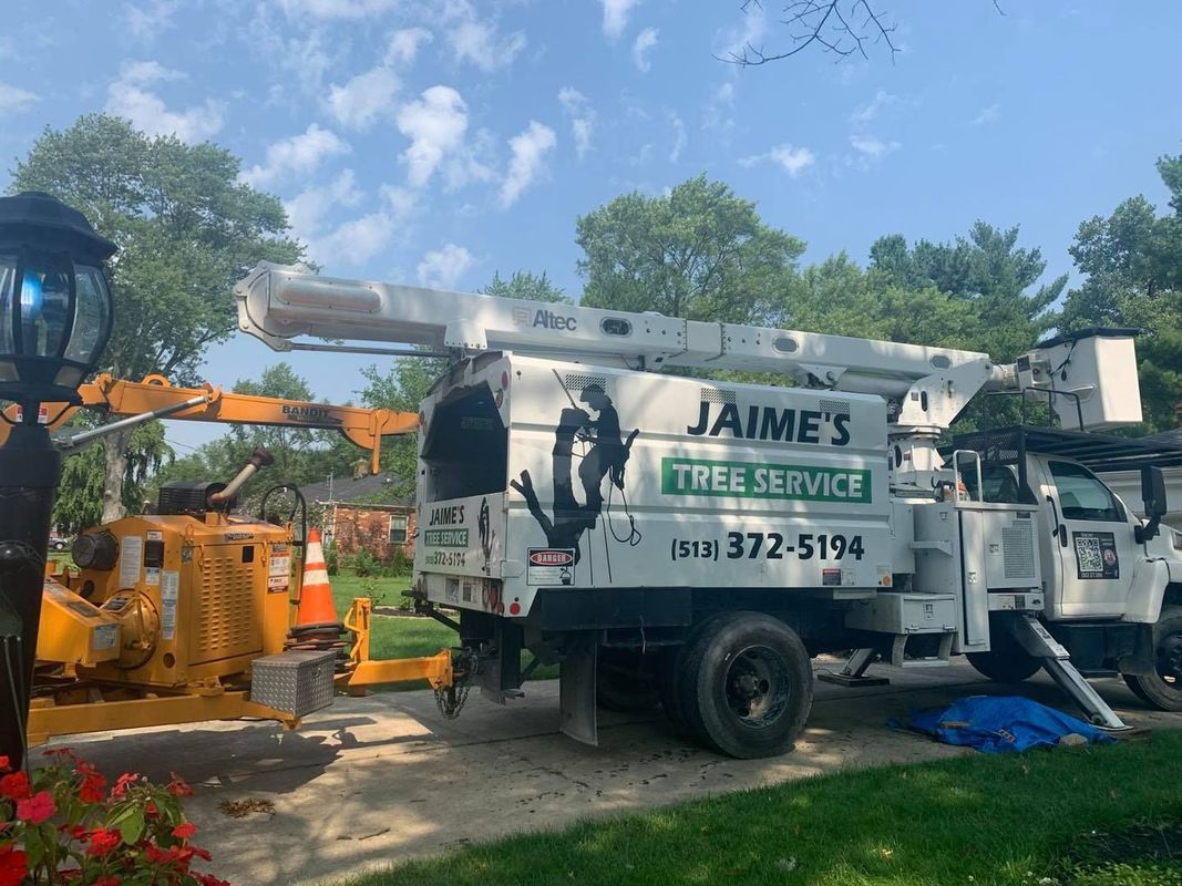 A Jaime’s Tree Service bucket truck parked in a driveway next to a yellow wood chipper on a sunny day.