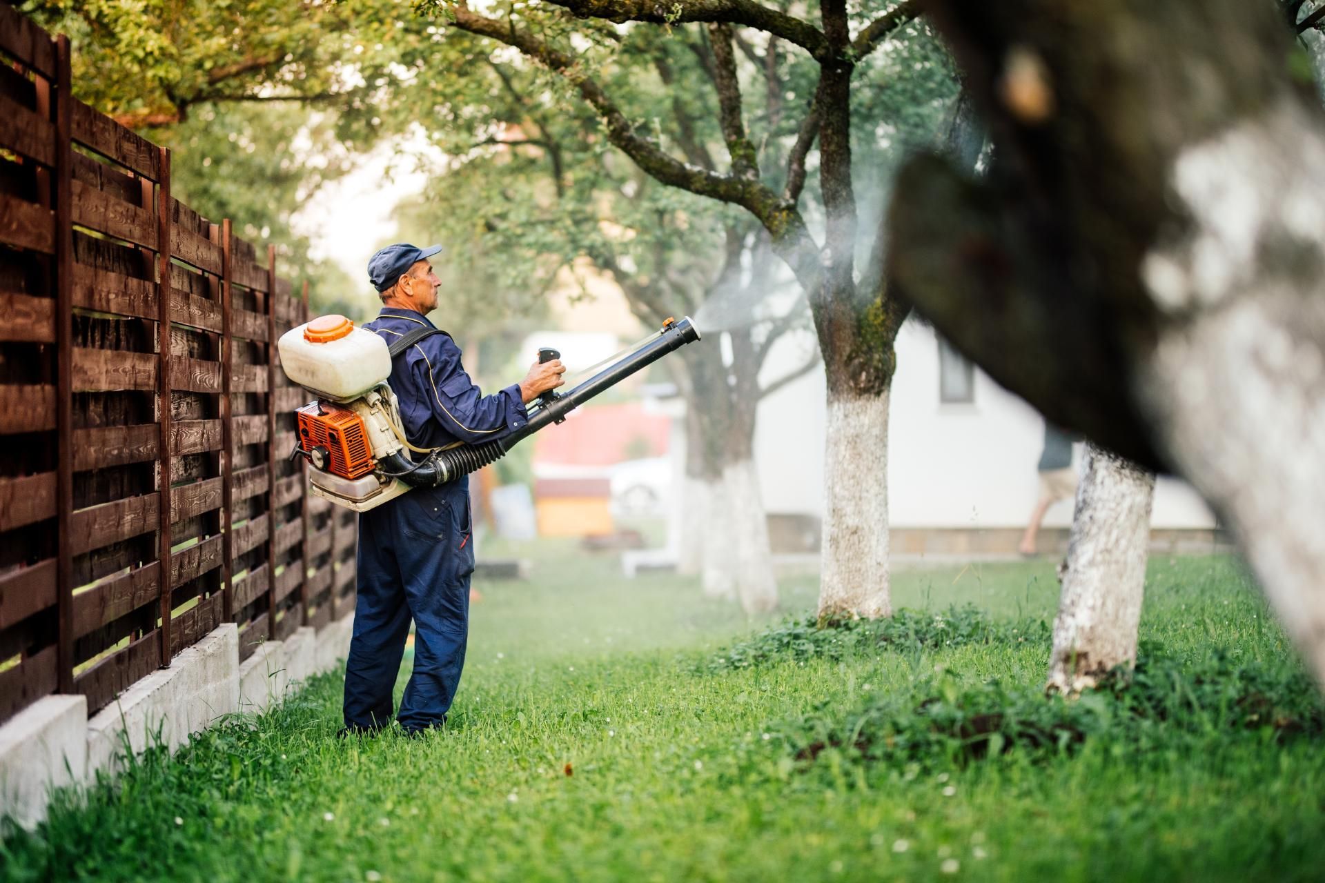A person in a blue uniform uses a backpack blower to spray mist into trees next to a wooden fence.