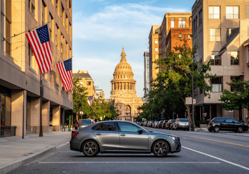 A gray car is parked on a downtown street with American flags displayed and the Texas State Capitol.