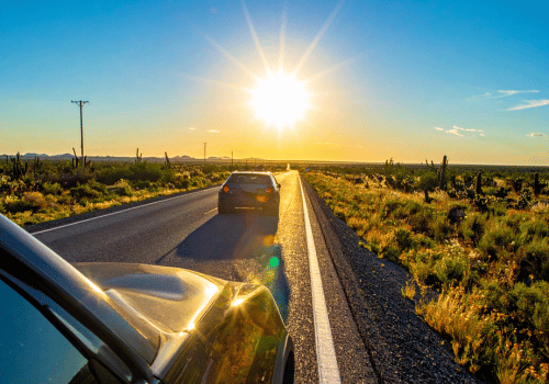 Two vehicles driving along a sunlit desert highway with cacti, open landscape, and clear Texas skies