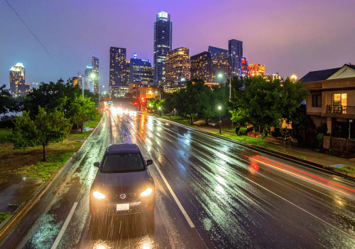 Car driving on a wet city street at night with reflections on the pavement and illuminated skyline.