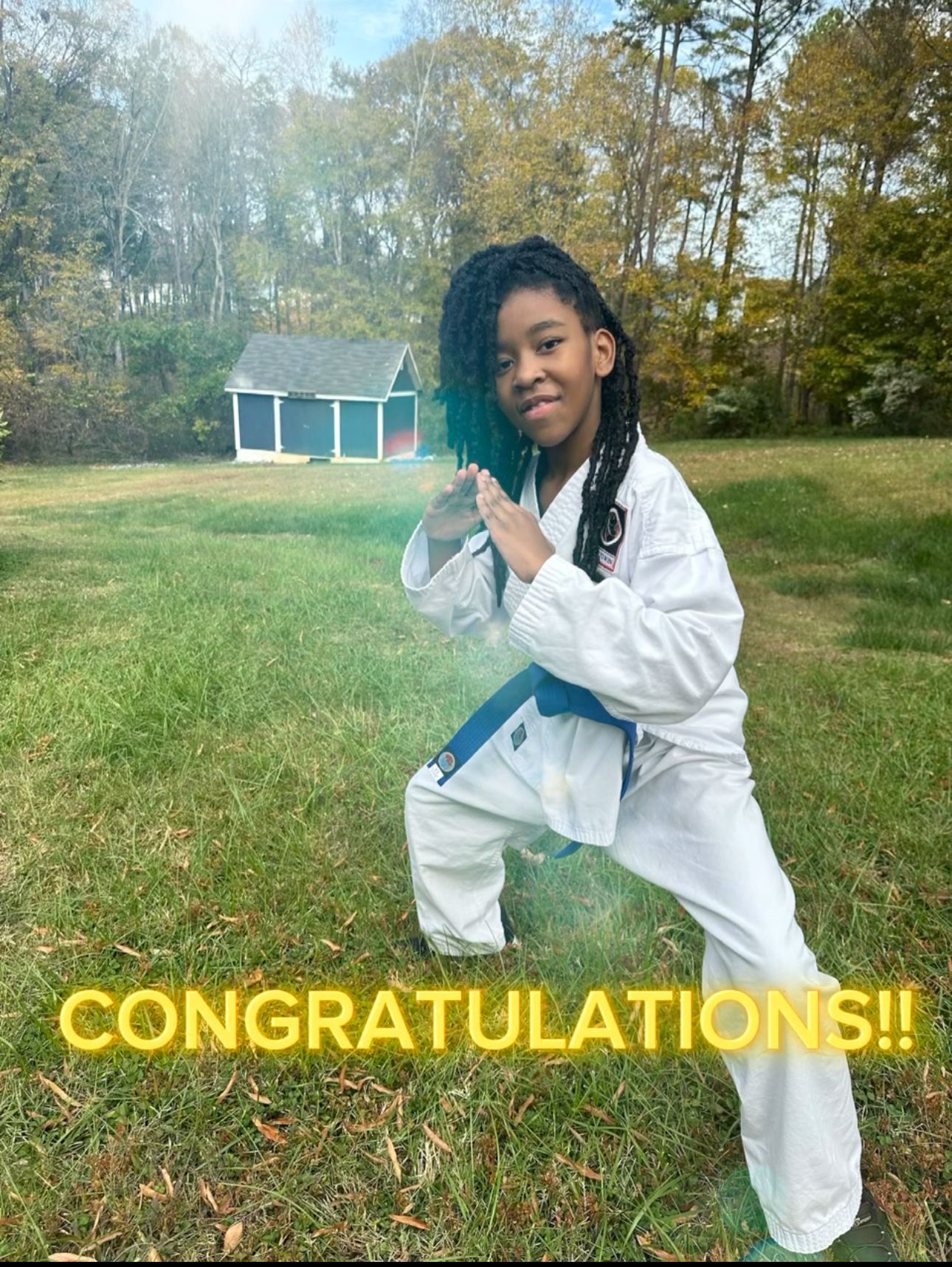 Young person in a karate uniform, blue belt, in a fighting stance on a grassy lawn with a small building in the background.