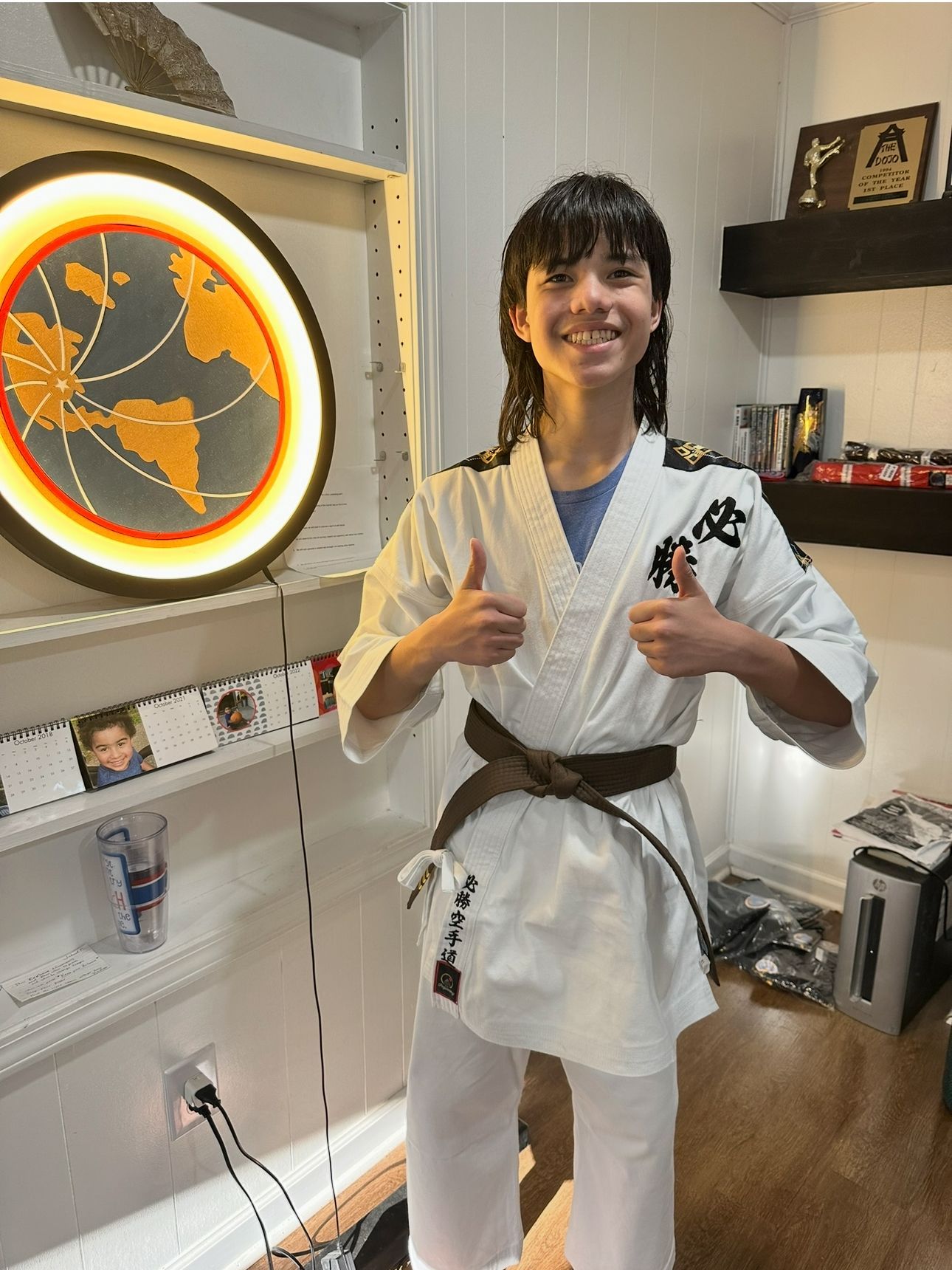Person in karate uniform with brown belt gives a thumbs up. Smiling, indoors with map and shelves.